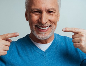 Man with white beard in blue sweater pointing to his smile